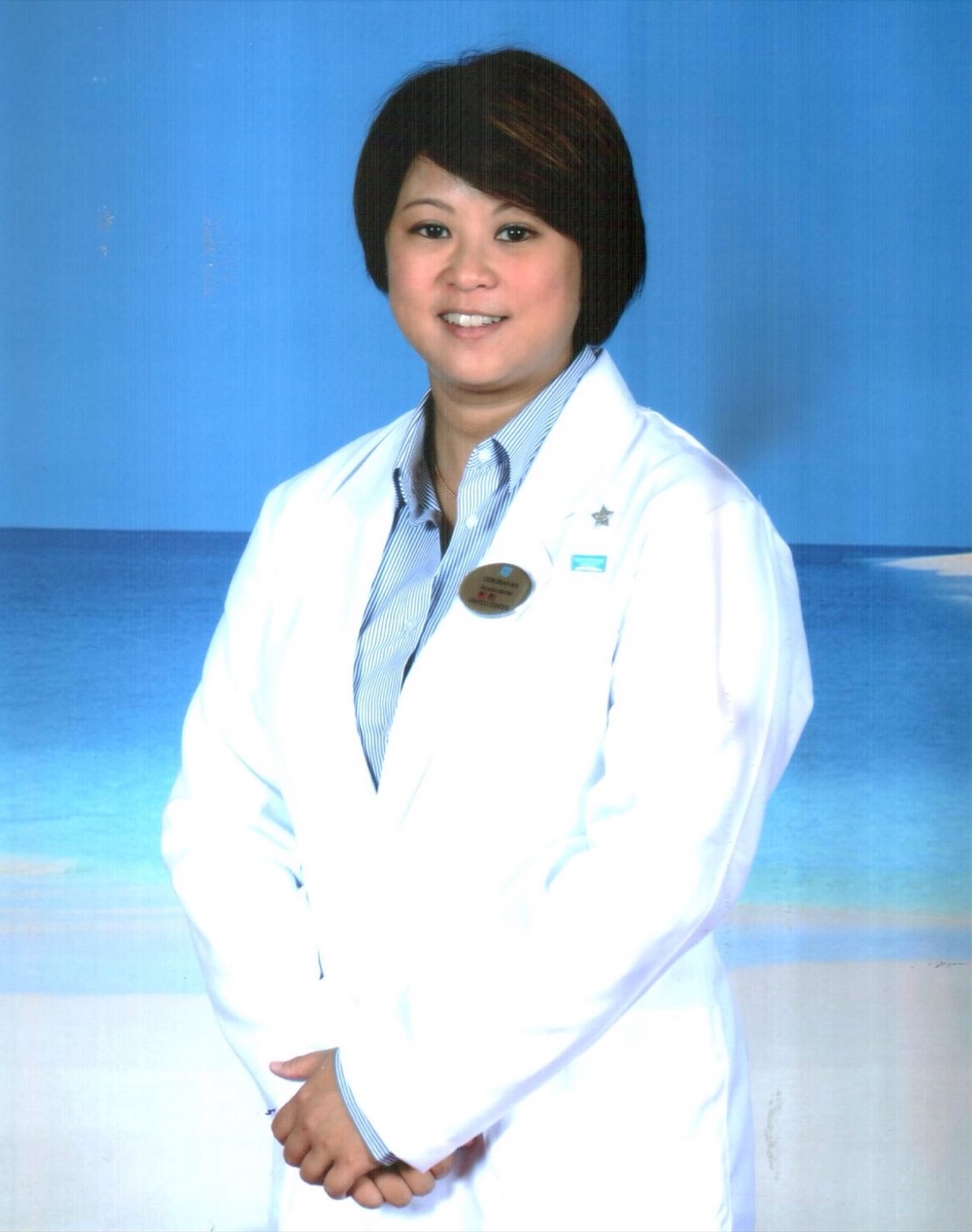 Dr Deb Ma in a white coat with a Norwegian Cruise Line name badge, photographed against the ship's painted blue backdrop.