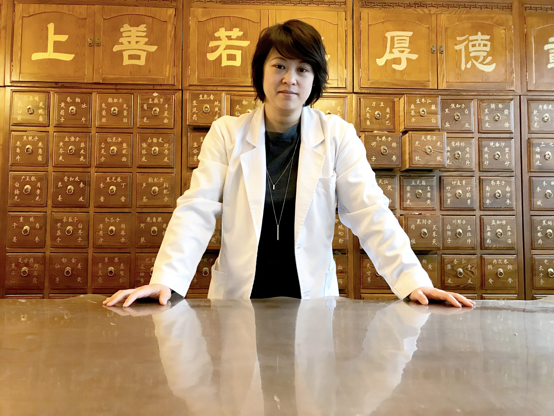 Dr Deborah Ma in a white coat, standing at a wall of Chinese herb drawers.