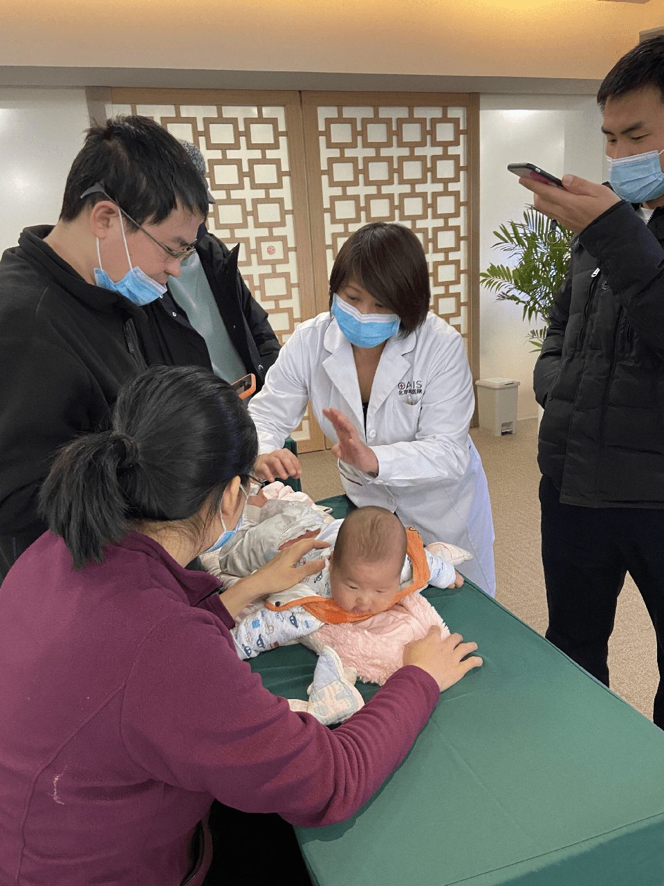 A masked clinician examines an infant on an exam table while family and colleagues look on, in a hospital training setting