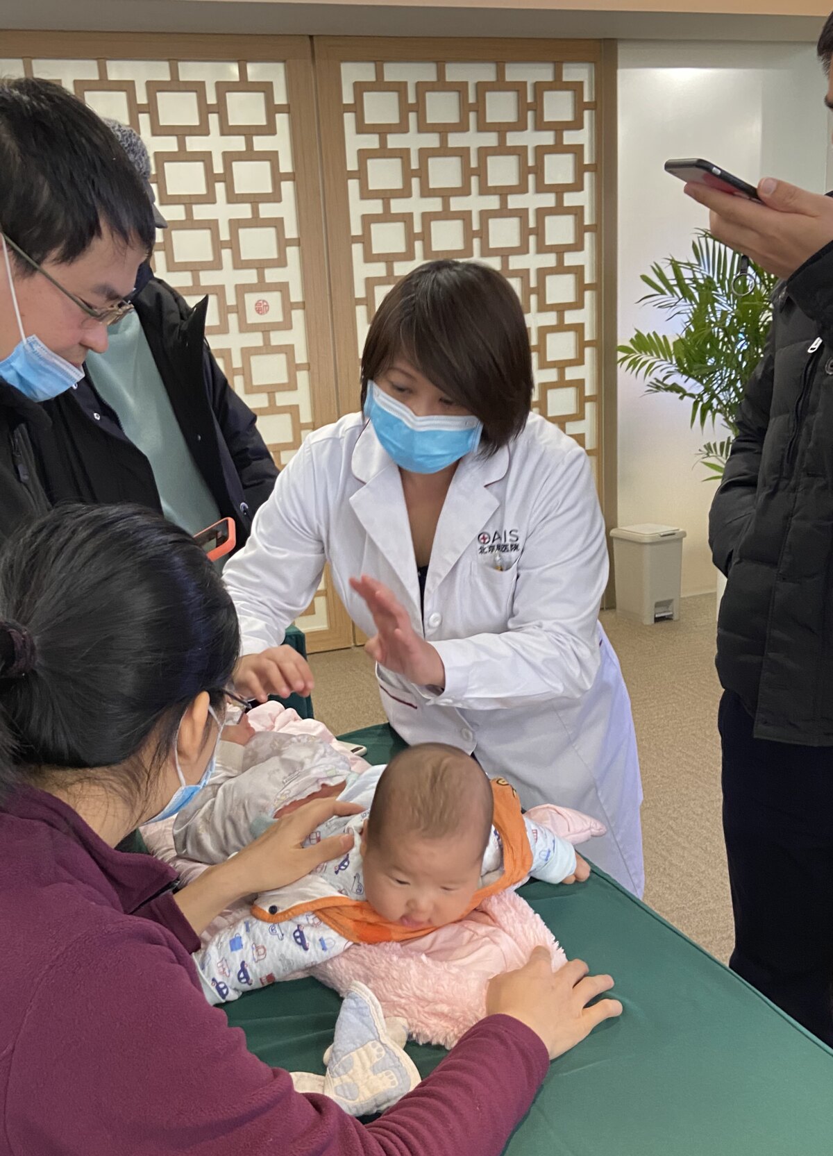 Dr Deb, in a white coat and mask, working on a baby lying on its front on a treatment table in a Beijing hospital, the mother holding the baby steady and two parents watching.